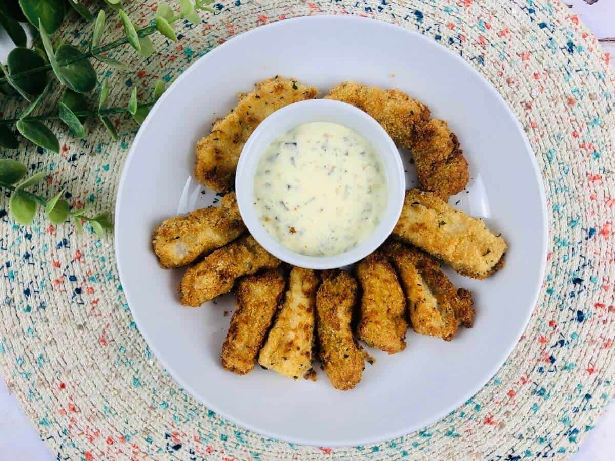 gluten free air fryer fish sticks on a white plate with a bowl of dip in the middle on top of a colorful placemat.