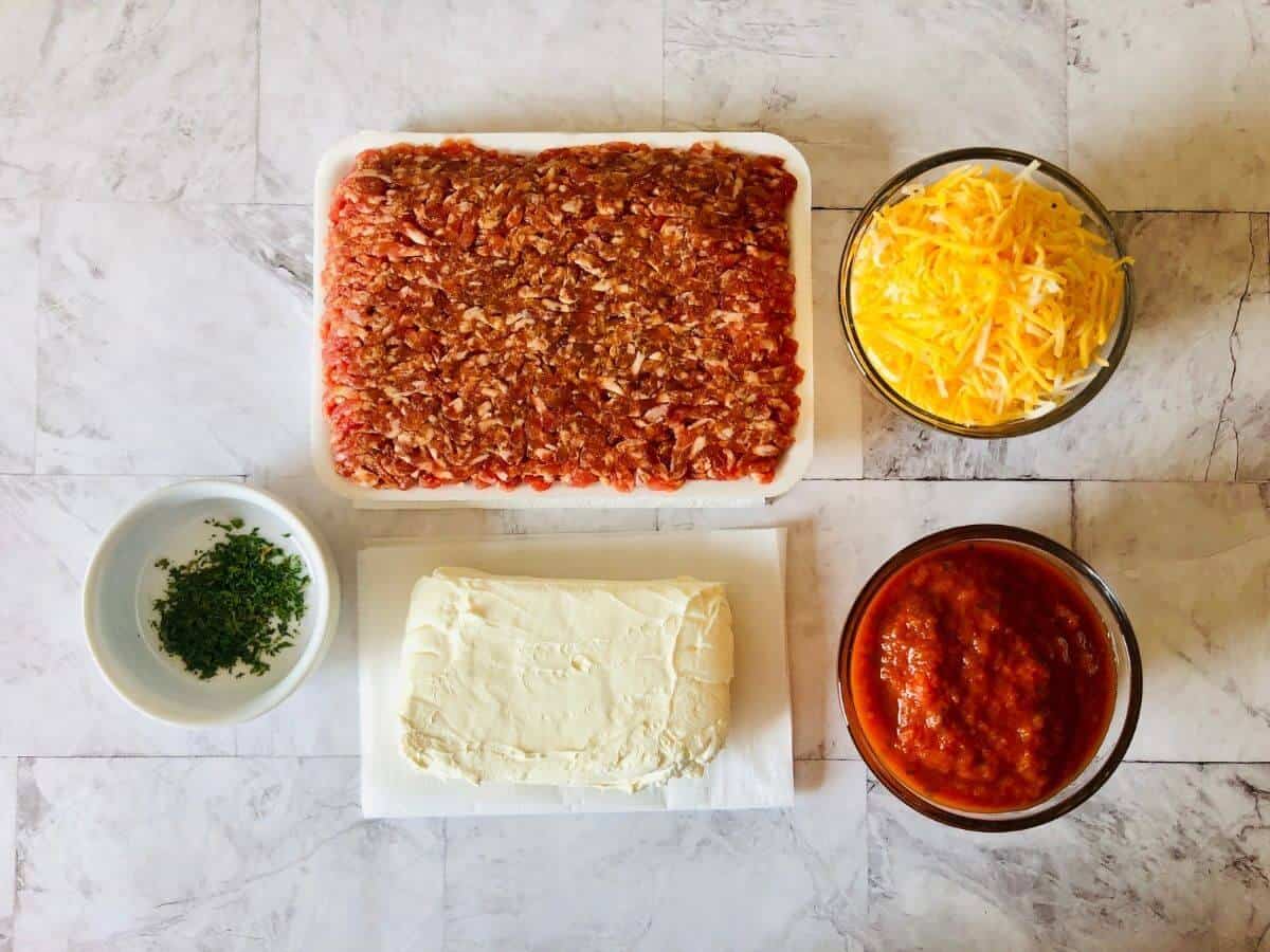 ingredients to make gluten free sausage balls including raw ground sausage, shredded cheese, herbs, cream cheese and red sauce all next to each other in bowls