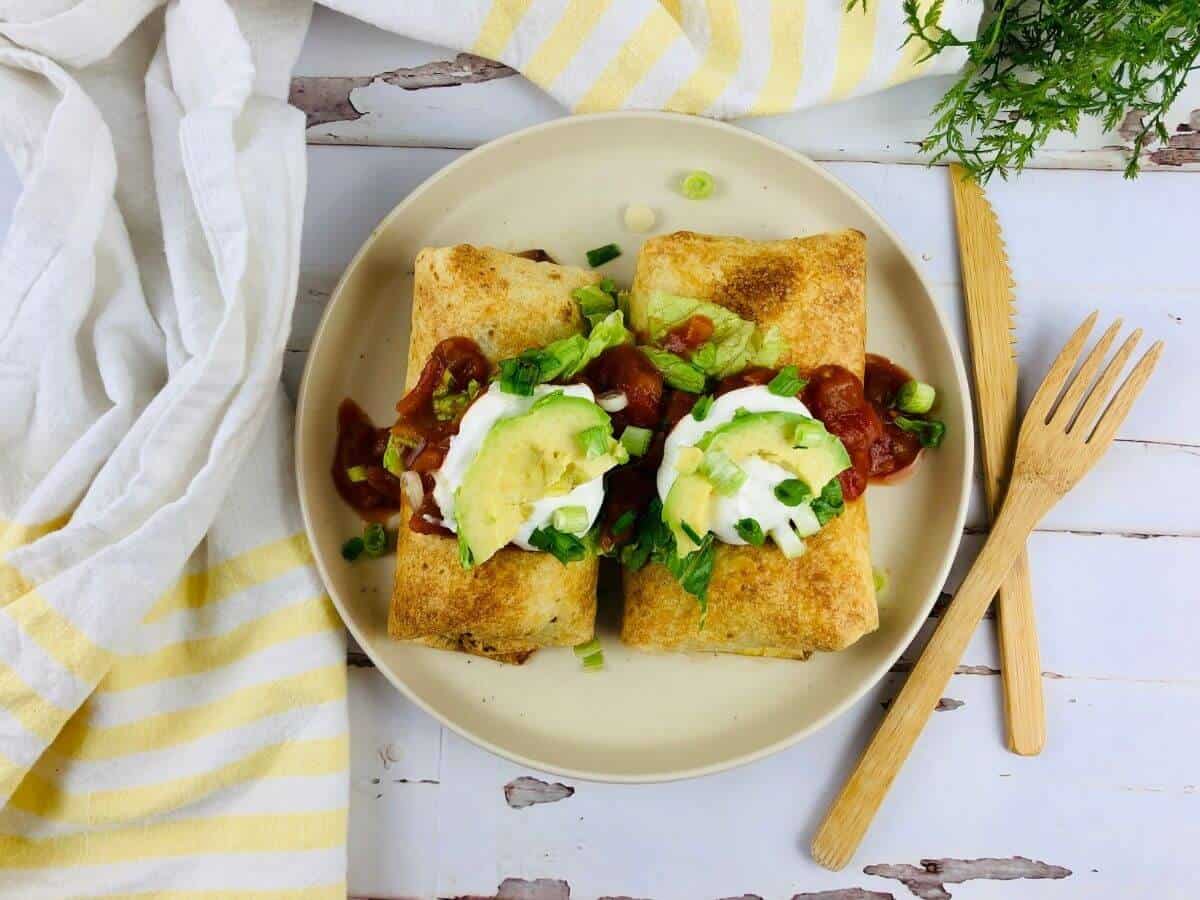 Two air fryer chimichangas on a plate loaded with toppings. Wooden fork and knife next to the plate.