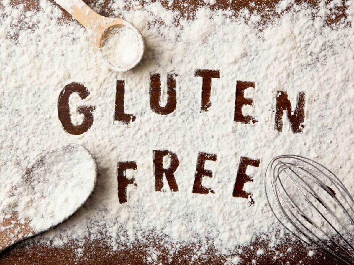 Flour covering a wooden table with "gluten free" written into it with cooking utensils on the side.