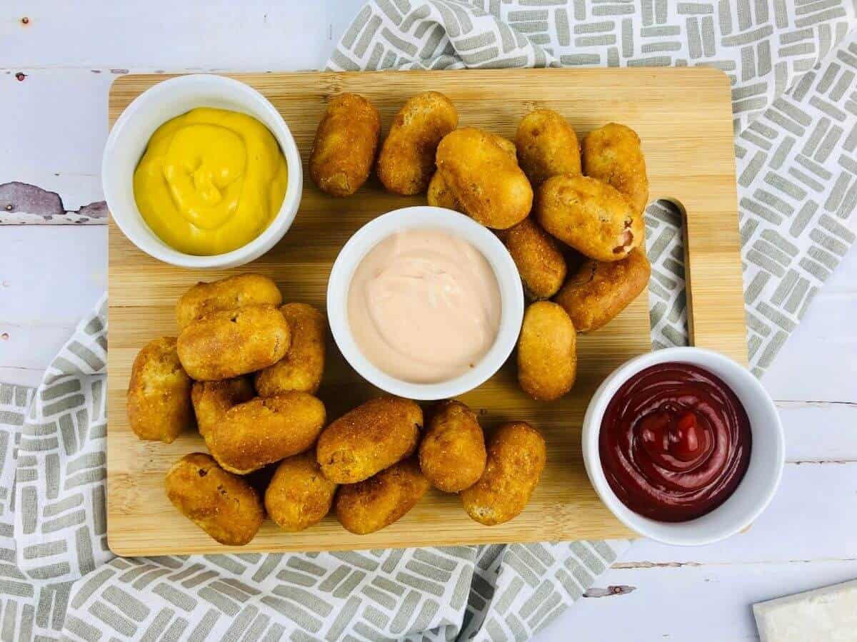 Wooden cutting board with gluten free corndogs, and 3 dip containers on top. Cutting board is on top of a decorative cloth napkin.