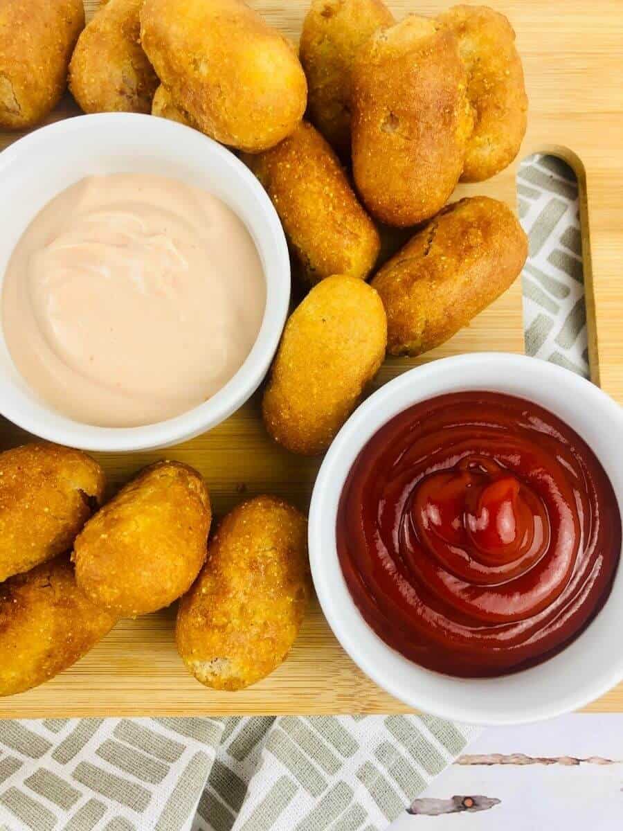 Close up vertical image of corn dogs on a wooden cutting board with 2 filled dip cups on the board.