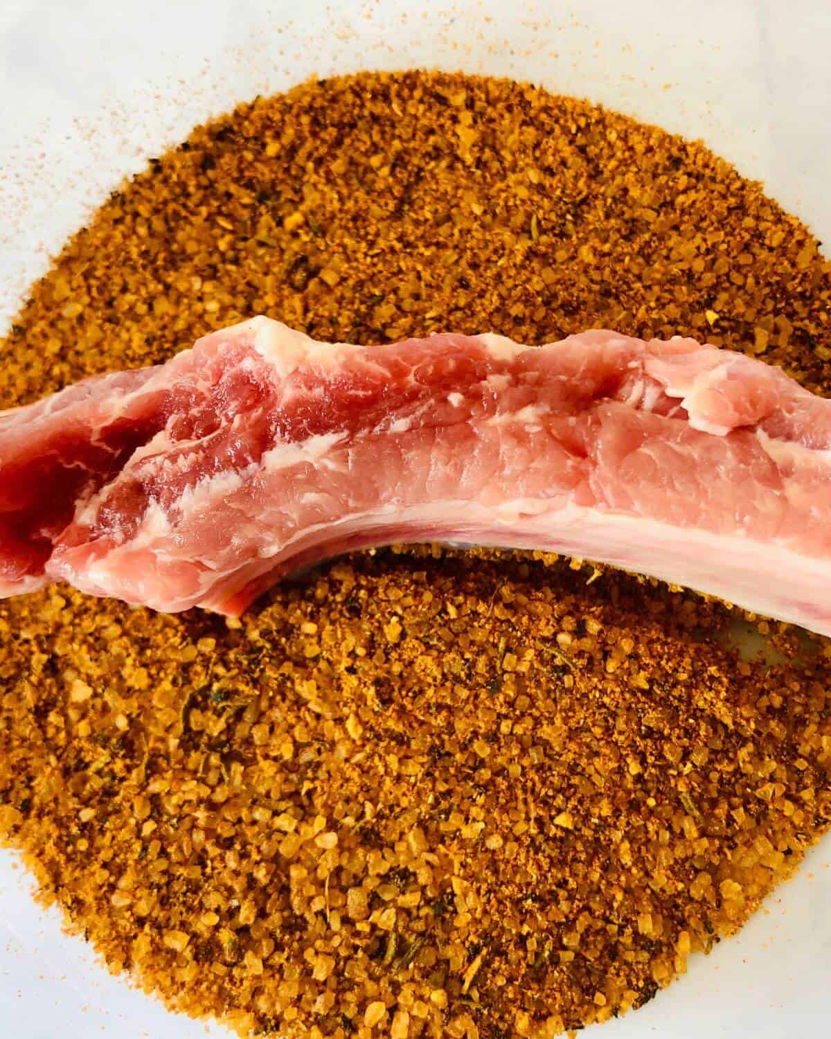 A raw pork rib placed in a bowl of dry rub seasoning.