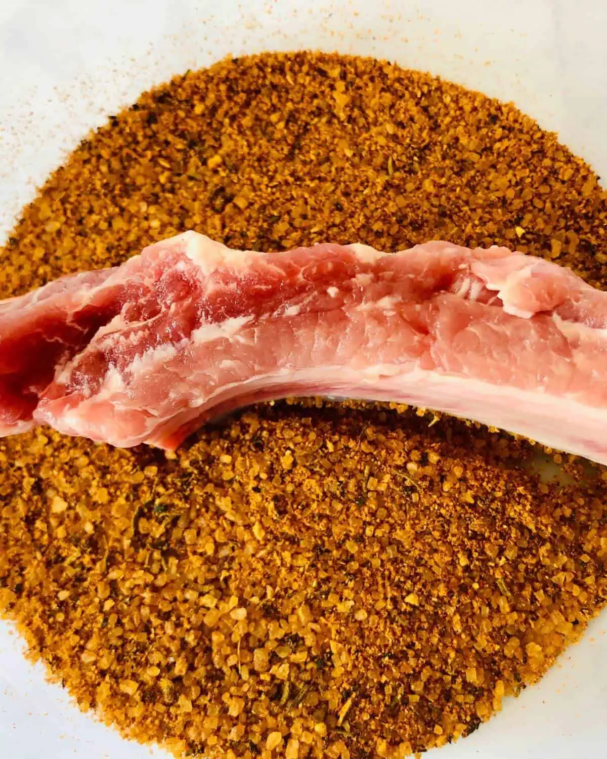 A raw pork rib placed in a bowl of dry rub seasoning.