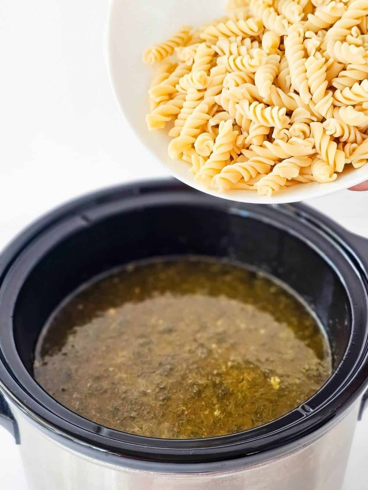 A bowl of cooked rotini pasta being poured into a slow cooker full of soup.