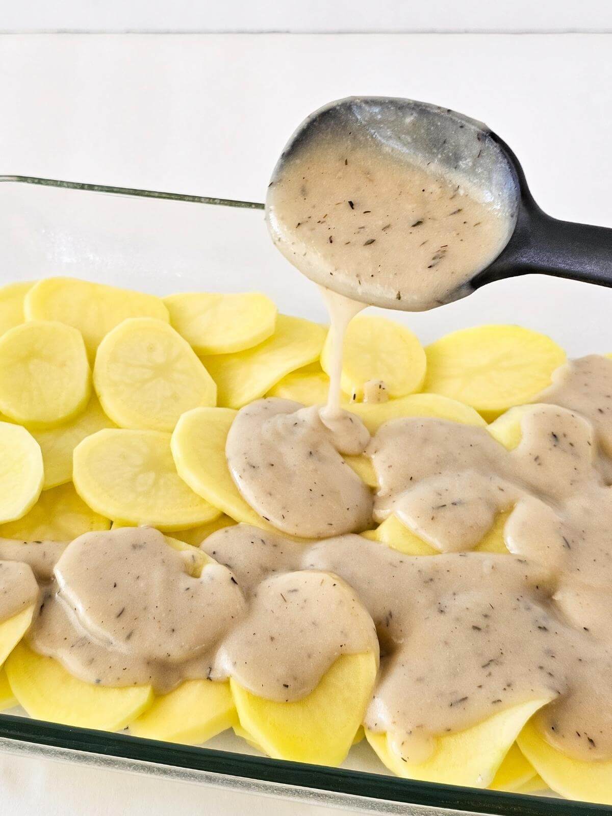 Cheese sauce being spooned onto sliced potatoes in a casserole dish.