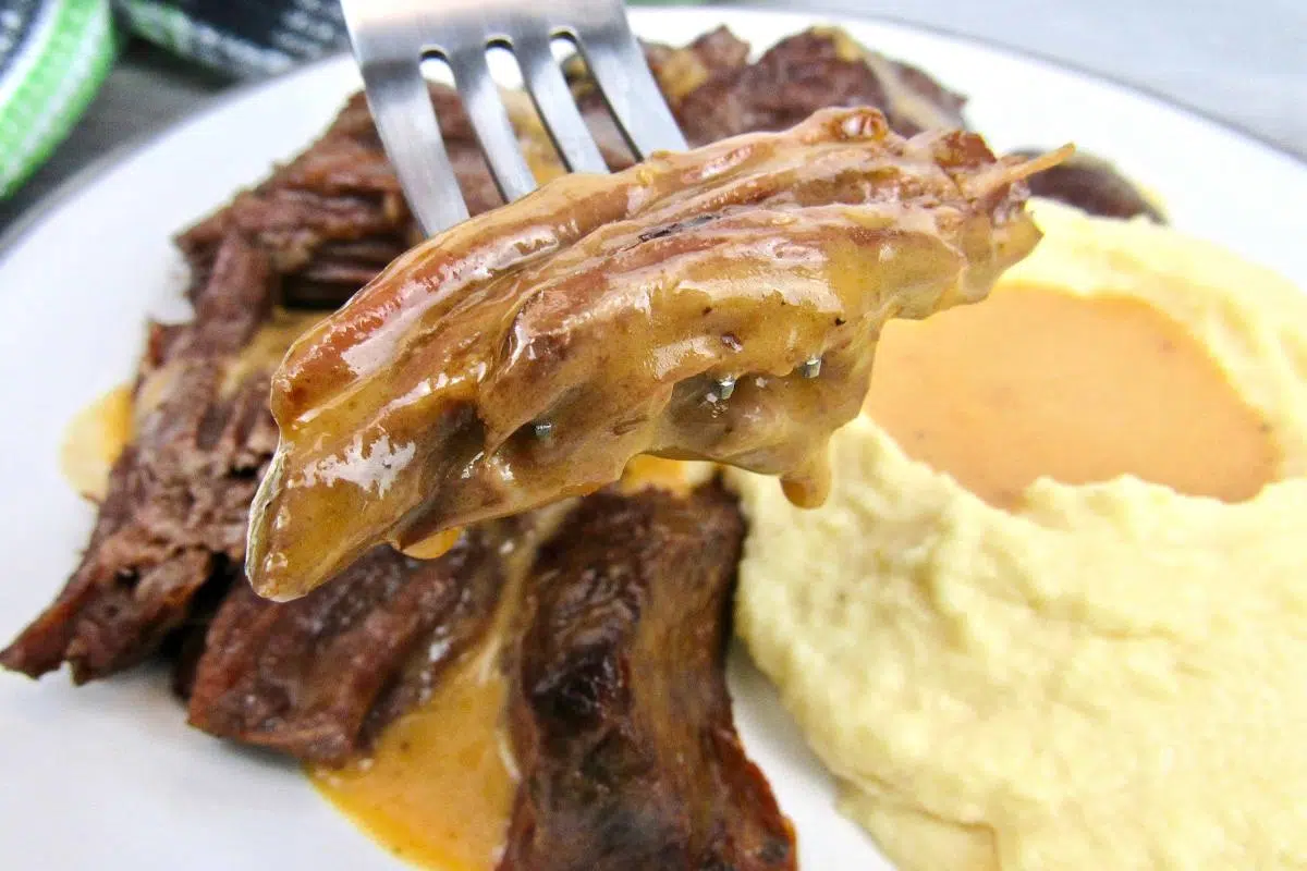 A fork picking up pot roast next to potatoes and gravy.