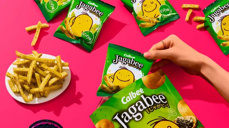 Hand holding a bag of Calbee Jagabee with some sticks on a plate on a pink background